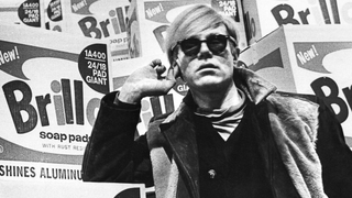 Artist Andy Warhol, a white man with blond hair wearing a black jacket, photographed in 1968 in front of boxes of Brillo