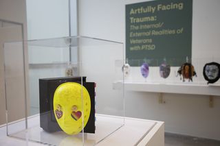 Colourful mask in display case at veteran PTSD art exhibition, with exhibition label in the background showing title, and a series of other masks on display