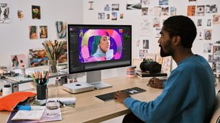 A person sits in front of an Apple Studio Display and a Mac mini on a desk.